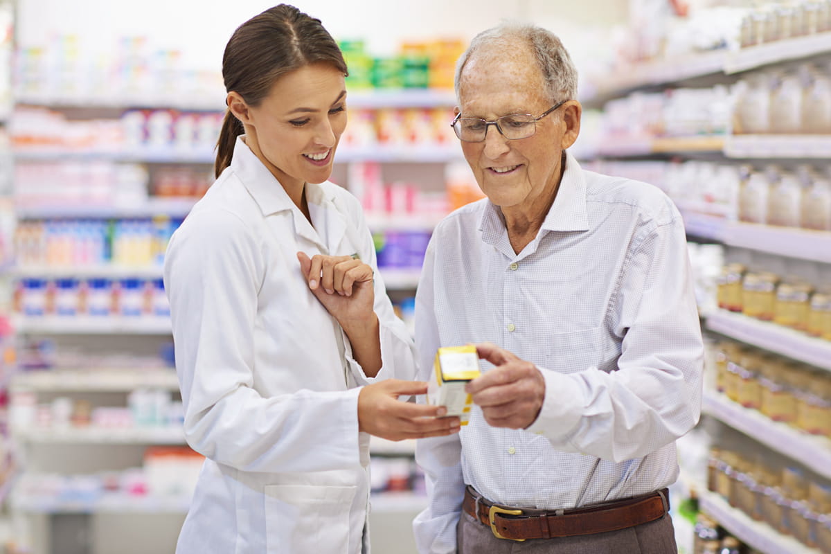 Senior man at pharmacy shows an OTC medication bottle to a pharmacist.